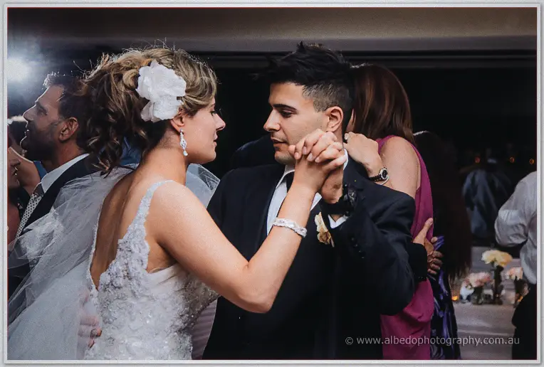 Bride in lace gown and white flower dancing with a young groomsman among the wedding guests at night