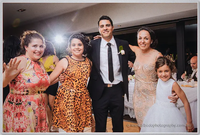 Groom in black tie posing with family and a little girl in white on the crowded dance floor