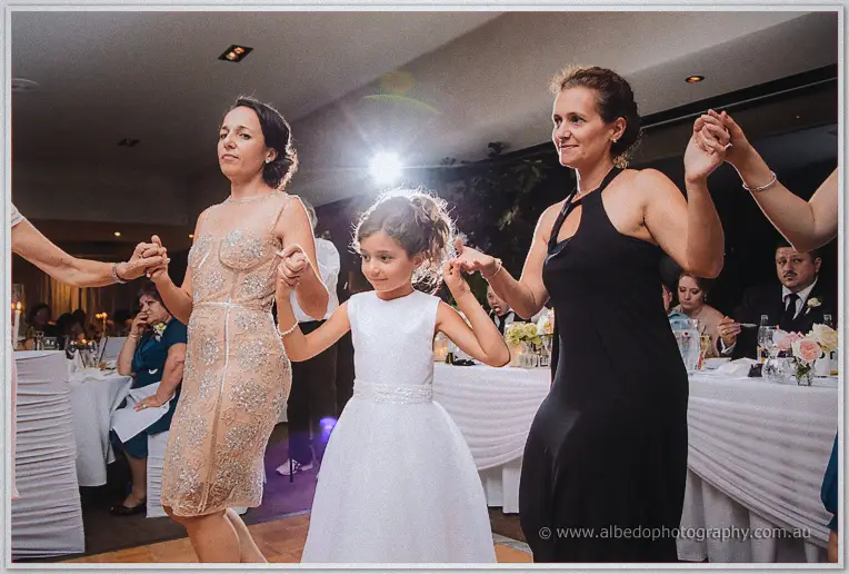 Flower girl in a white dress dancing hand in hand with two smiling women in sequinned and black gowns