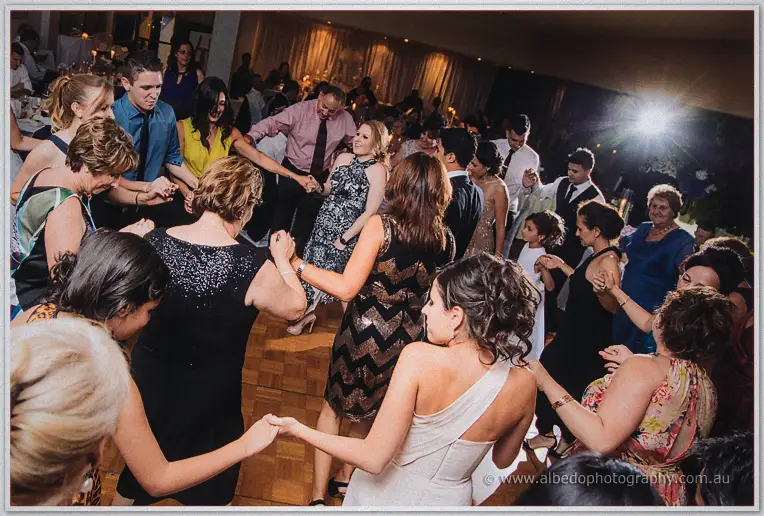 Bride in a one-shoulder gown dancing in a hand-linked circle of women on the busy parquetry floor