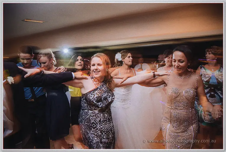 Bride with a white hair flower dancing between guests in patterned and gold sequin gowns, arms outstretched