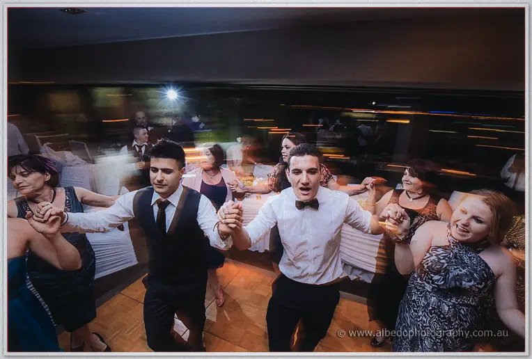 Two young men in vest and bow tie dancing in a guest conga line, streaked lights behind them