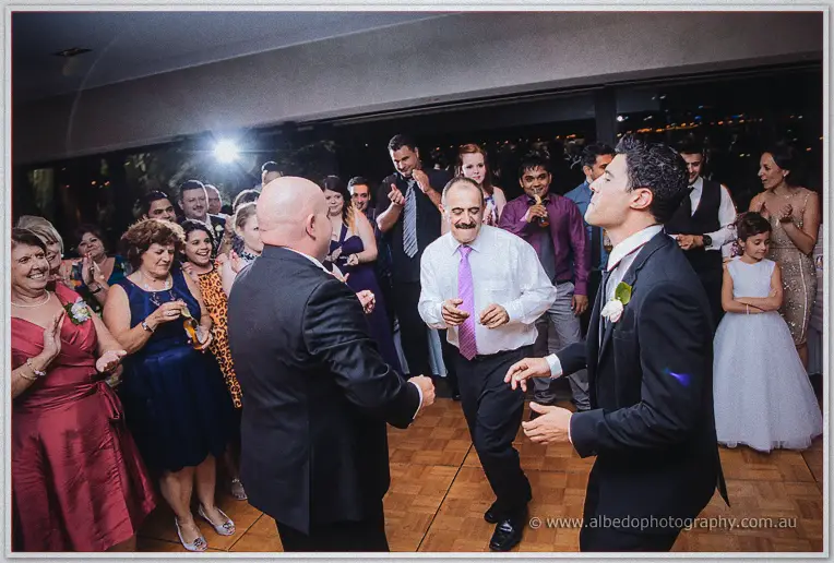 Mustached man in a pink tie dancing centre circle as the groom and guests clap around him