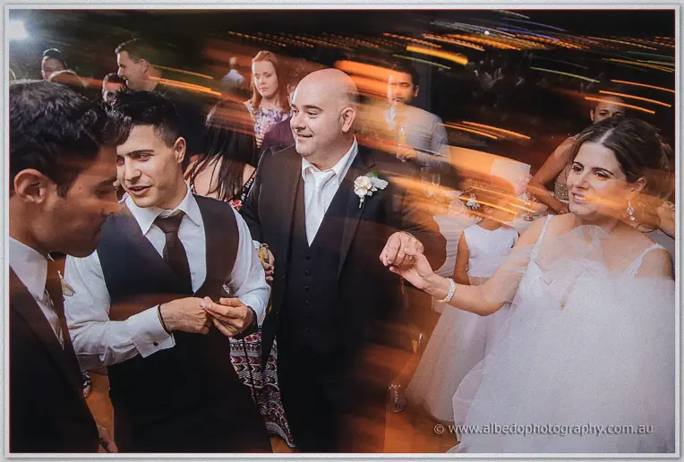 Groom with a cream boutonniere dancing in a blur of spinning guests and warm streaked lights at night