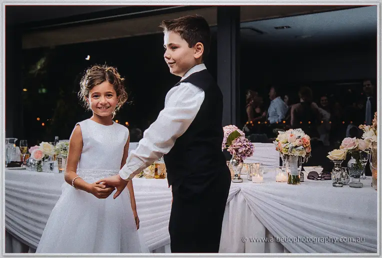 Page boy in black vest holding hands with a smiling flower girl in white before the candlelit bridal table