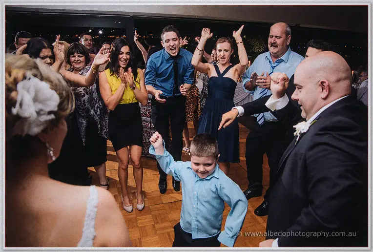 Page boy dancing with fist raised in the middle of a cheering guest circle, bride watching from the side
