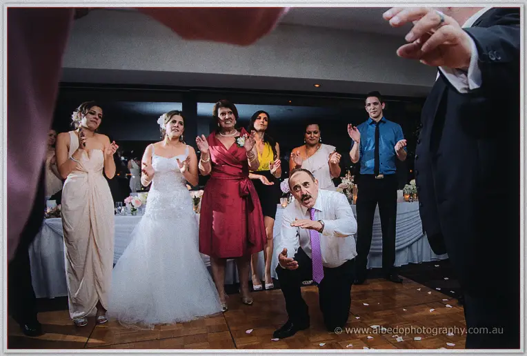 Mustached man in a lilac tie crouching low as the bride and guests dance and clap behind him
