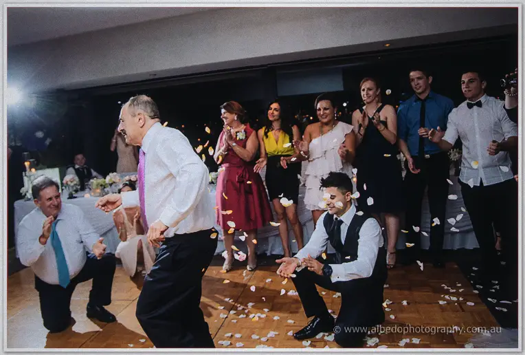 Men in ties crouch and whirl in a circle dance as rose petals scatter across the parquetry floor