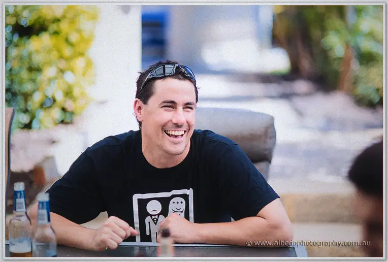 A laughing groom in a black tee waits with drinks at an outdoor table on the morning of the wedding