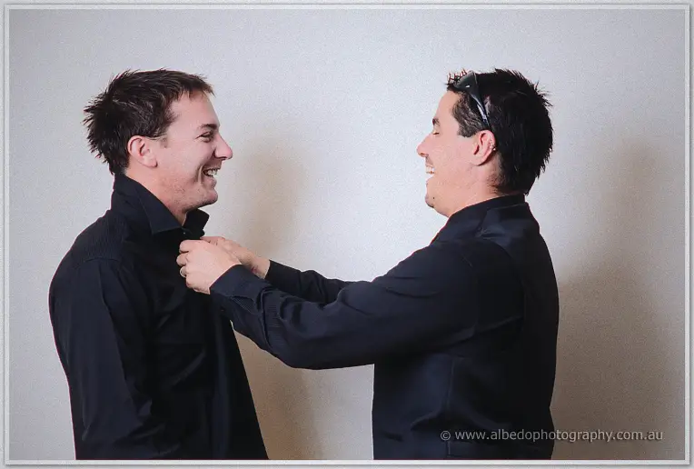 Two groomsmen in black shirts laugh as one fixes the other's collar against a plain wall