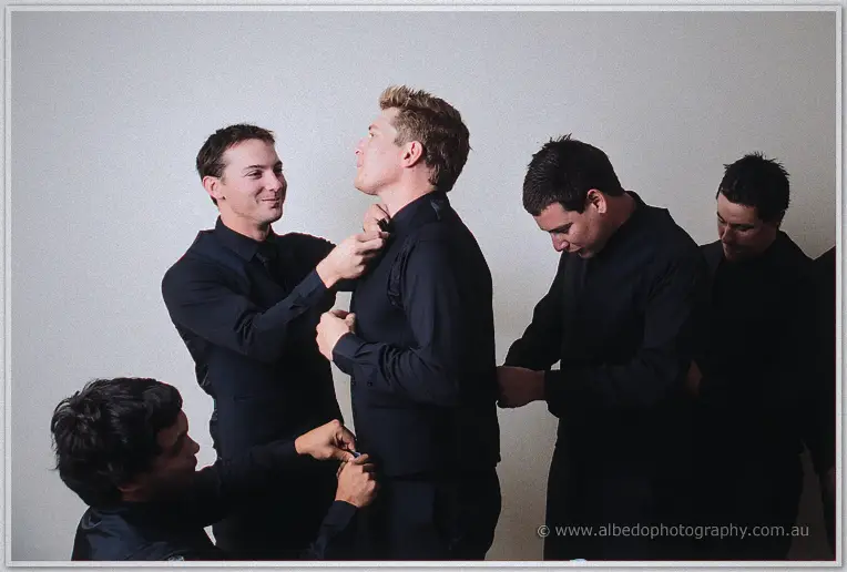 Groomsmen in matching black help Matt with his vest, buttons and collar before the ceremony