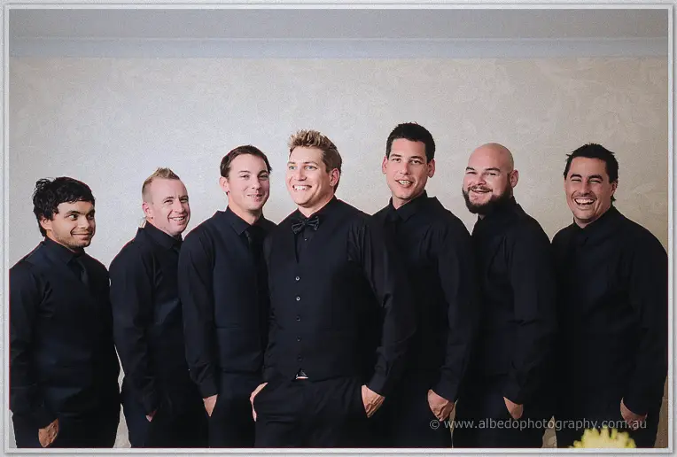 Matt stands with his six groomsmen, all in black shirts and vests, grinning at the camera