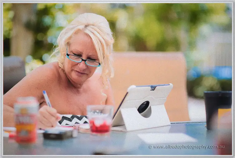 A blonde wedding guest in reading glasses writes notes beside an iPad on an outdoor table