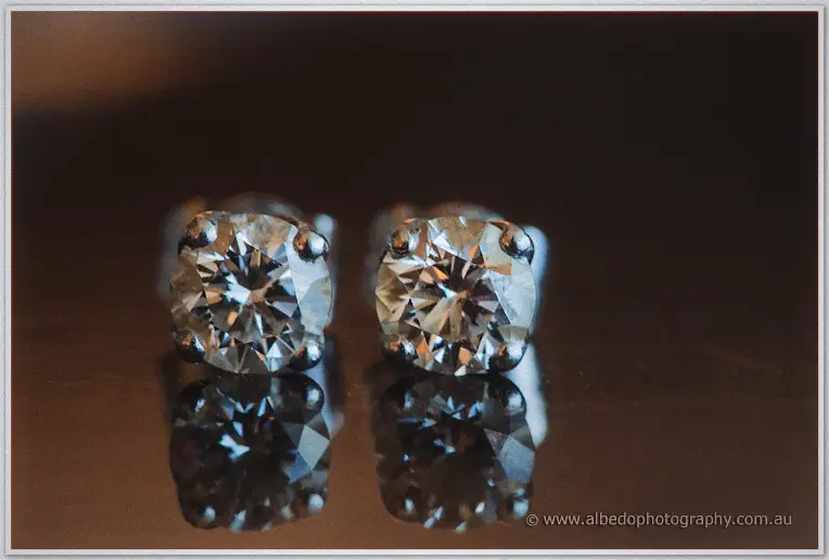 Close-up of Haley's diamond stud earrings sparkling against a dark reflective surface in warm morning light