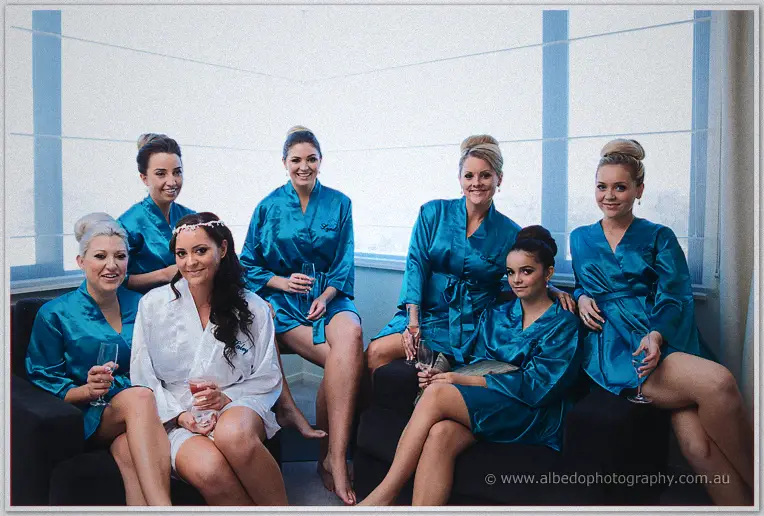 Haley and her bridesmaids in teal satin robes toast with champagne by a bright hotel window