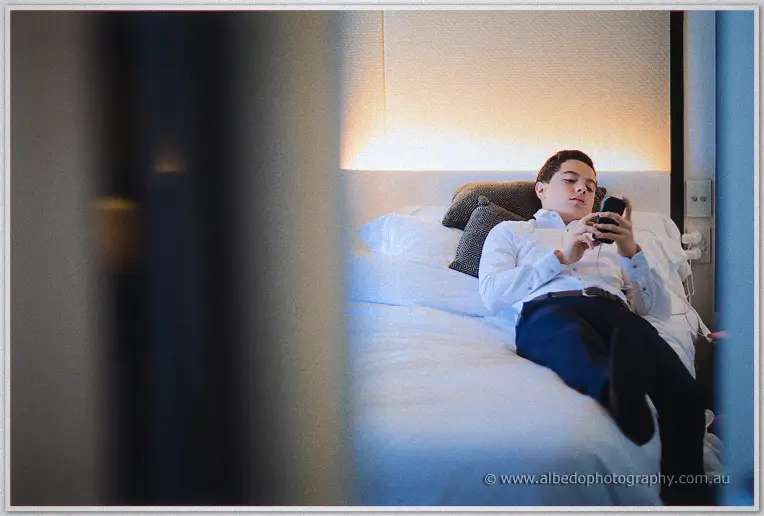 A page boy in white shirt and navy trousers lounges on a hotel bed playing on his phone