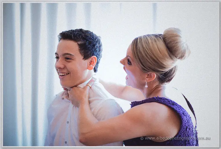 A woman in a purple dress fixes a smiling page boy's collar in soft window light