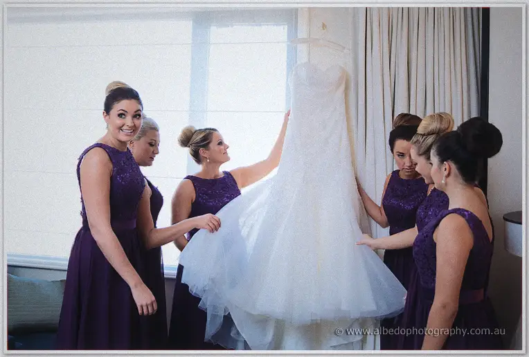 Bridesmaids in purple lace dresses gather around Haley's white gown hanging in window light