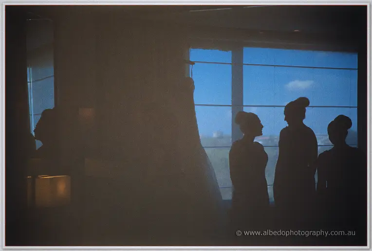 Bridesmaids stand silhouetted by a blue window beside Haley's hanging gown before the ceremony