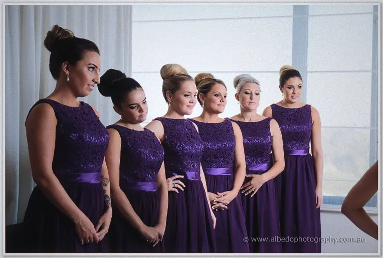 Six bridesmaids in matching purple lace dresses line up by the window light before the Caversham ceremony