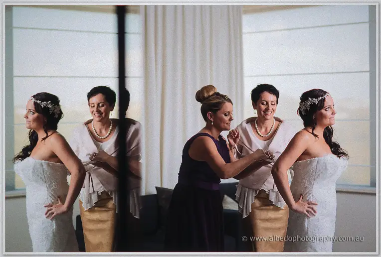Bride Haley stands hand on hip as her mother and bridesmaid adjust her bracelet beside bright hotel windows