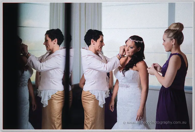 Haley's mother fastens an earring on her daughter while a bridesmaid in purple looks on by soft window light