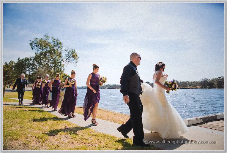 Haley leads her purple-dressed bridesmaids along a riverside path toward the jetty in bright sunshine
