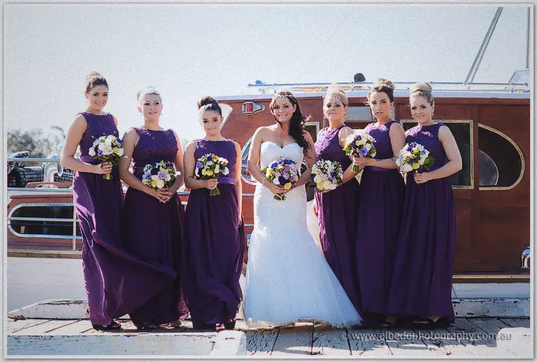 Haley and her six bridesmaids pose with bouquets on a weathered timber jetty before a classic wooden cruiser