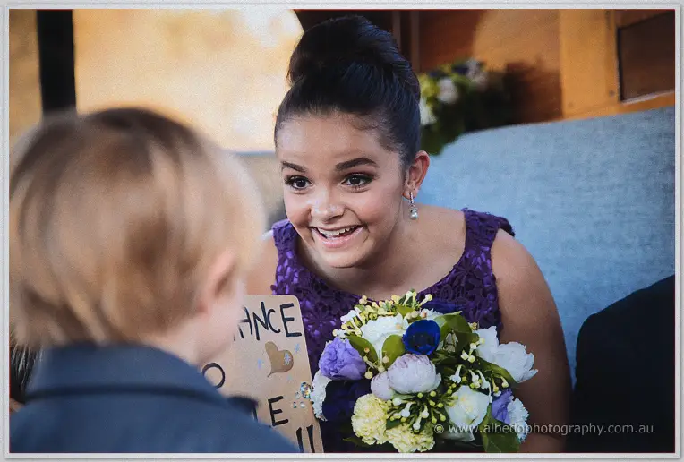A junior bridesmaid in purple beams at a small boy holding a handwritten sign among the floral bouquets