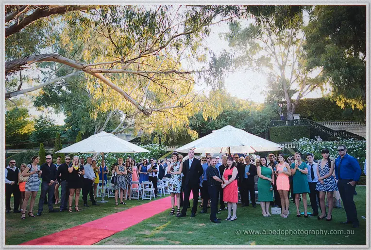 Guests gather along the red carpet aisle under white umbrellas as golden light filters through the gums