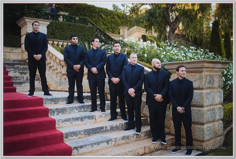 Groomsmen in black suits line Caversham's limestone steps beside the red carpet under fragrant garden white
