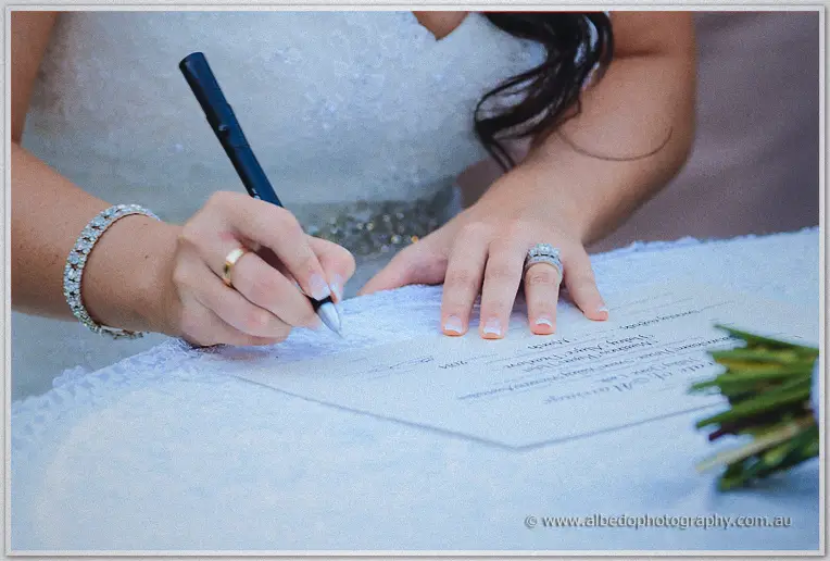 Haley signs the marriage certificate, a close-up of her hands with gold rings on the lace-covered table.