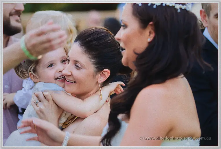 A beaming flower girl in white hugs one of the wedding guests as Haley watches the sweet moment beside them.
