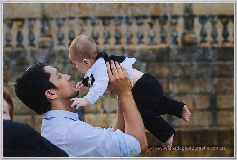 A groomsman lifts a baby in suspenders and tiny black trousers into the air by the limestone fountain wall.