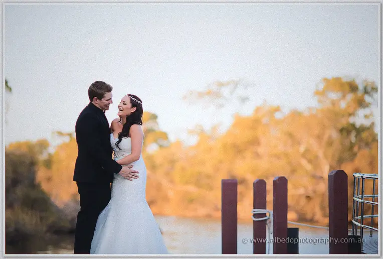 Haley laughs in Matt's arms on the riverside jetty, golden trees glowing across the water behind them.