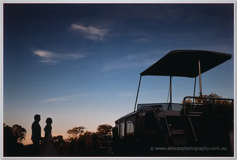 Haley and Matt stand silhouetted against a deep blue dusk sky beside the canopied boat at the river's edge.