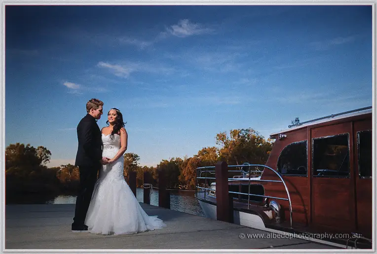 Haley and Matt stand together on the jetty beside the polished timber launch under a bright blue evening sky.