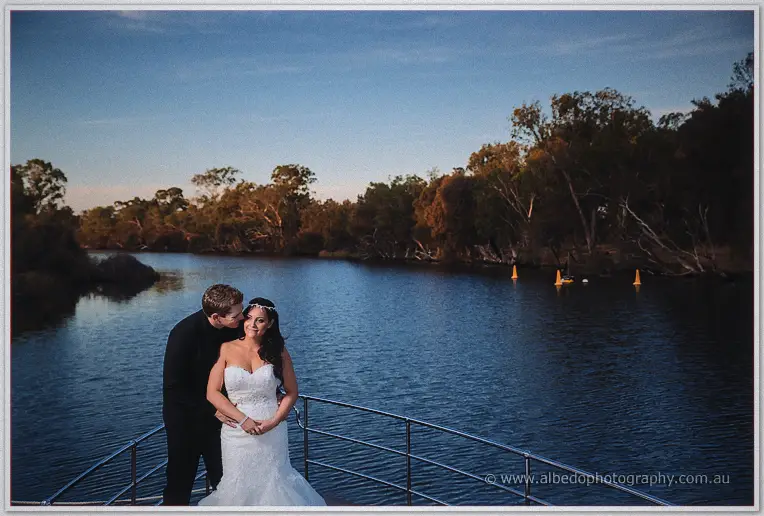 Matt kisses Haley's cheek on the boat's bow deck as still river water and gum trees stretch out behind them.