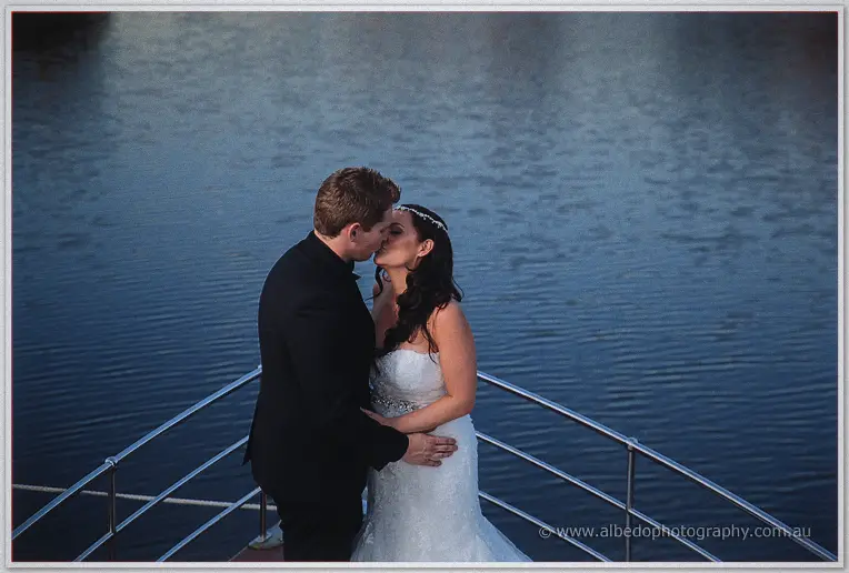 Haley and Matt share a kiss on the boat's bow deck, dusk-blue river water rippling behind them.