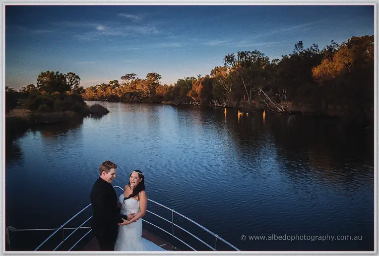 Haley and Matt laugh together at the boat's railing, the tree-lined river glowing at sunset behind them.
