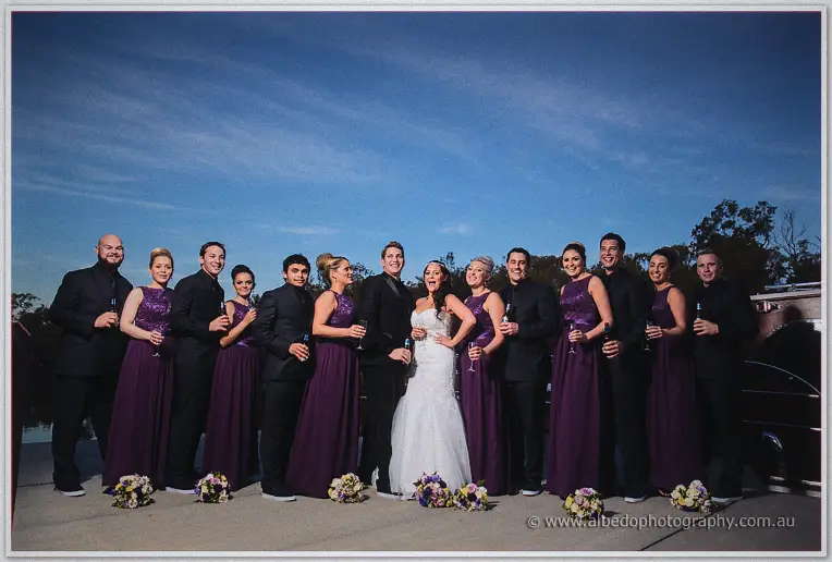 The bridal party raises champagne bottles and glasses at dusk, bouquets lined along the ground at their feet.