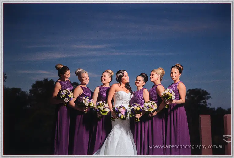 Haley stands with her bridesmaids in purple lace gowns holding purple and cream bouquets at evening.