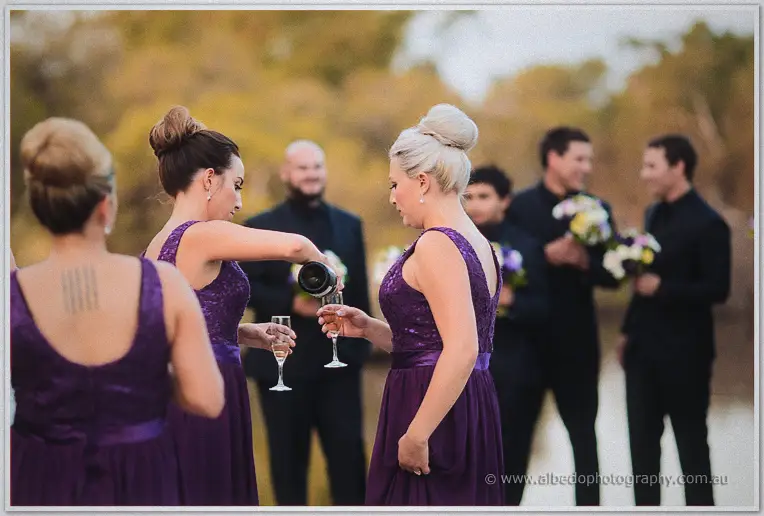 Bridesmaids in purple sequin gowns pour champagne on the riverbank while groomsmen wait with bouquets.