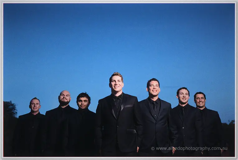 Matt and his groomsmen stand shoulder to shoulder in black suits against a clear twilight sky.