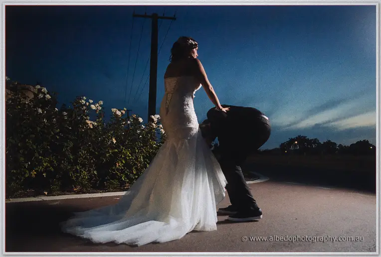 Matt bows to adjust Haley's flowing train beside white rose bushes in the last light of evening.