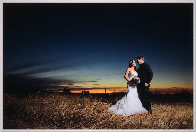 Haley and Matt stand close in a dry golden paddock, her bouquet of purple blooms catching the last daylight.