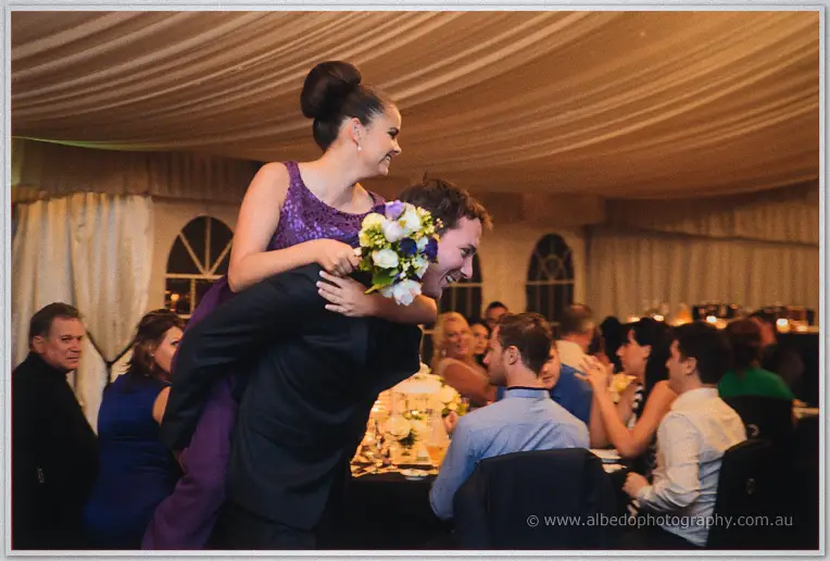 A groomsman piggybacks a laughing bridesmaid in purple sequins past seated wedding guests in the marquee.