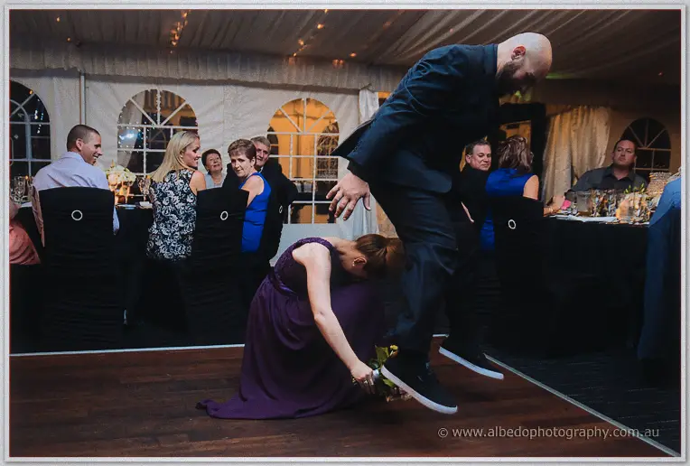 A groomsman leaps over a bridesmaid stooping for the bouquet on the marquee dance floor.