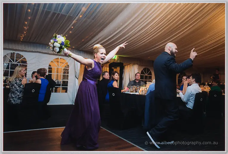 A bridesmaid in purple celebrates catching the bouquet with both arms flung wide beside a dancing groomsman.