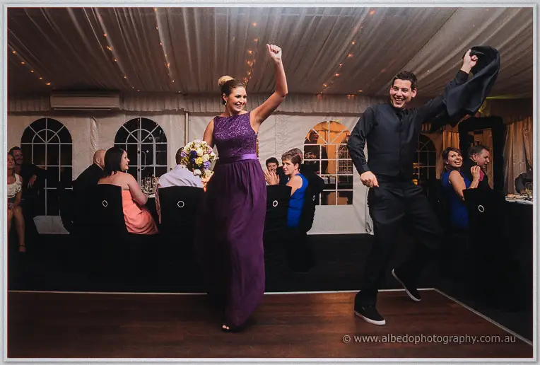 A bridesmaid in purple and a groomsman in black dance with raised fists on the marquee's wooden dance floor.